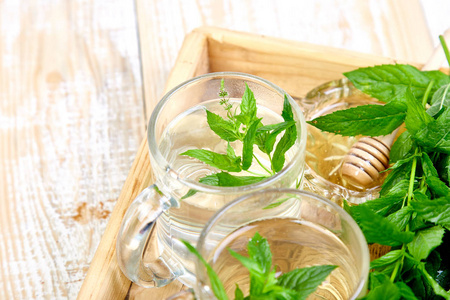 热的草药薄荷茶饮料在玻璃杯或杯子上的木制托盘。 蜂蜜和新鲜花园薄荷叶复制空间。
