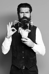 英俊的胡须男子与五颜六色的管长胡子和胡子有时尚的头发在严重的脸上举行着酒精鸡尾酒在老式绒面革皮革背心蓝色工作室背景