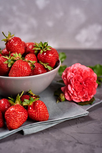 红色成熟草莓在陶瓷碗上的亚麻餐巾纸和粉红色玫瑰花在灰色混凝土背景，复制空间。健康食品概念，静物