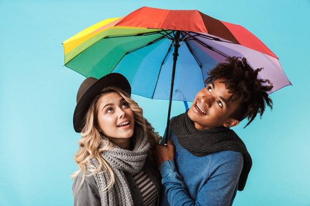 戴着围巾的年轻多种族夫妇微笑着站在蓝色背景上，带着雨伞