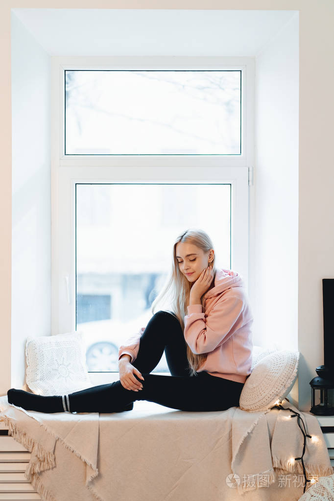可爱的女孩坐在窗台上,抱着枕头,在室内思考和做梦.