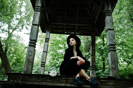 的女孩都戴着黑色的红色嘴唇和帽子。 哥特式戏剧女人坐在森林的木拱上。