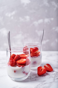 两杯健康酸奶，新鲜切片草莓和勺子在白色大理石桌背景复制空间。 健康早餐食品概念