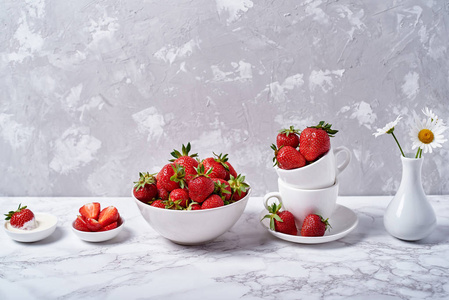 成熟的有机草莓放在白色陶瓷碗里，奶油和洋甘菊花放在灰色混凝土背景的花瓶里，复制空间。健康食品理念，静物