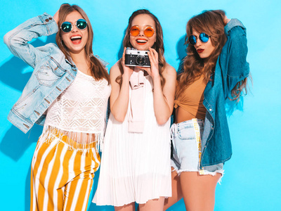三个年轻美丽的微笑时髦女孩穿着时髦的夏季休闲服装和太阳镜。 无忧无虑的女人在蓝墙附近摆姿势。 拍摄复古相机
