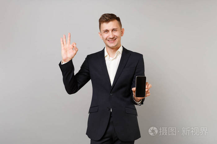 有吸引力的年轻商人穿着黑色西装,展示着ok手势,手持手机,灰色背景上