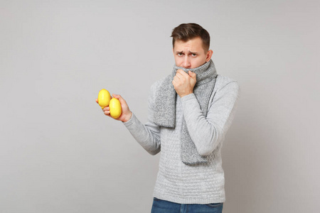 心烦意乱的年轻人穿着灰色毛衣，用围巾盖住嘴巴，在工作室里用灰色背景隔离的柠檬。 健康生活方式病态疾病治疗寒冷季节的概念。 模拟复