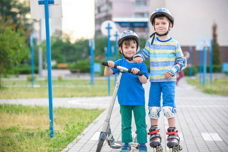 两个男孩在溜冰鞋和他的兄弟在滑板车包裹在公园。 孩子们戴着安全轮滑的保护垫。 儿童积极户外运动。