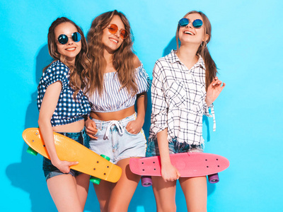 三个年轻时尚微笑美丽的女孩与五颜六色的硬币滑板。夏天，穿着时髦衬衫的女人在工作室里摆在蓝墙附近。积极的模特喜欢戴太阳镜