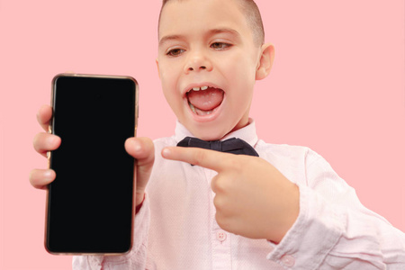 室内肖像画，吸引人的小男孩被隔离在粉红色的背景上，拿着空白的智能手机，对着相机微笑，显示屏幕，感到快乐和惊讶。人类情感，面部表情