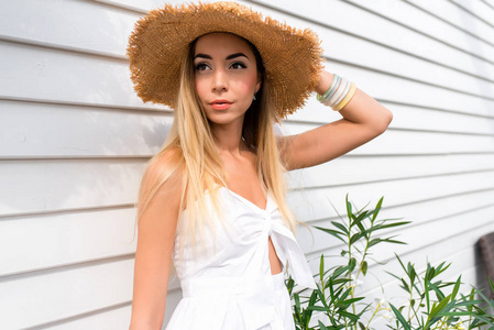 美丽的女孩在草帽在木白色墙壁背景。女人在衣服与晒黑的皮肤和日常化妆。夏天在阳光门廊上