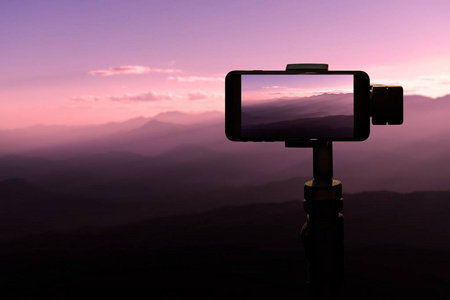 旅行者使用智能手机在三脚架上拍照在日落山自然背景。