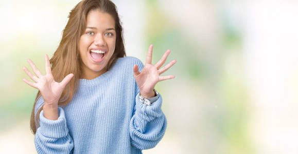 年轻漂亮的黑发女人穿着蓝色的冬季毛衣，在孤立的背景下庆祝疯狂和惊讶的成功，举起手臂，睁开眼睛尖叫兴奋。赢家概念