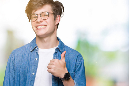 年轻的英俊男子戴着眼镜，在孤立的背景上，用手做快乐的拇指向上的手势。 赞许的表情看着相机显示成功。