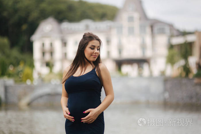 美丽的年轻开朗的孕妇站在喷泉的背景.怀孕6个月了她把手放在肚子上