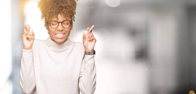 美丽的年轻非洲裔美国妇女戴着眼镜，在孤立的背景上微笑着交叉手指，充满希望和眼睛紧闭。 运气和迷信的概念。