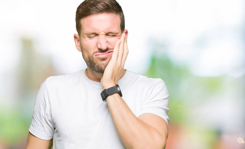 英俊的男人穿着休闲的白色T恤，由于牙痛或牙齿疾病，用手摸嘴，表情痛苦。 牙医的概念。