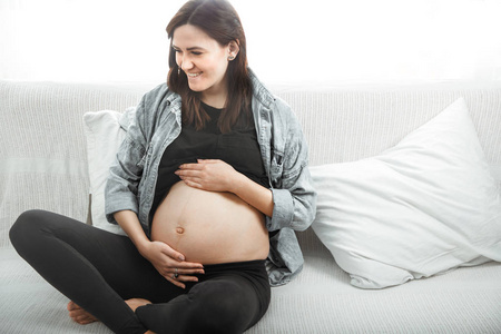 年轻漂亮的孕妇坐在沙发上靠窗怀孕的观念和对孩子的期待