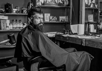 戴着黑斗篷的大胡子坐在镜前的美发椅上。男子与胡子客户的时髦理发店。理发店概念。时髦与胡子等待理发和理发