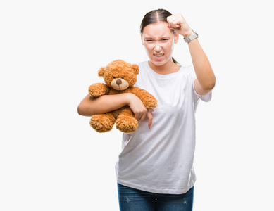 年轻的白种人女人抱着泰迪熊，在孤立的背景下烦恼和沮丧，愤怒，疯狂地大喊大叫，举起手愤怒的概念