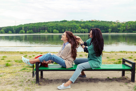 两个漂亮的女朋友玩得开心，放松和微笑。 深色头发做头发到红色，而坐在长凳上，背景是可怕的绿色景观。