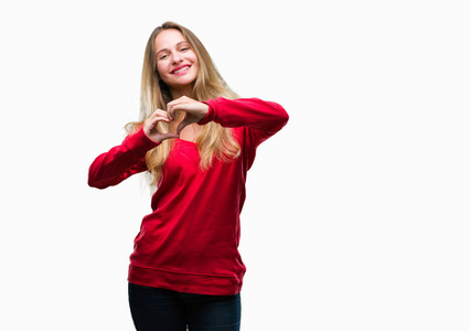年轻漂亮的金发女人穿着红色毛衣，在孤立的背景上微笑，在爱中显示心脏的象征和形状与手。 浪漫的概念。