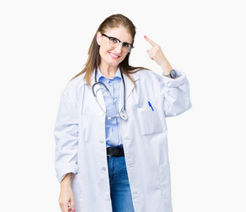 中年成熟的医生女人穿着医用外套，在孤立的背景下微笑着用一根手指指着头，伟大的想法或美好的记忆