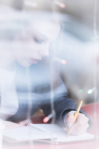 一个有吸引力的年轻记者女孩的肖像与笔和记事本在咖啡馆背后的展示。通过窗户玻璃反射的非对比度视图