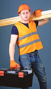 男子头盔, 硬帽子持有工具箱和木梁, 灰色背景。木匠, 木工, 劳动者, 建筑工人在平静的脸上扛着木梁。木工概念