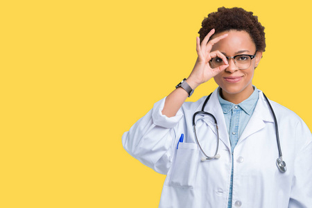 年轻的非裔美国医生女人穿着医用外套，在孤立的背景下做着OK的手势，用手微笑的眼睛透过手指看着幸福的脸。