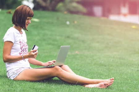 女性网上购物的概念。绿色公园里的女人配着现代化的笔记本电脑。巴厘岛