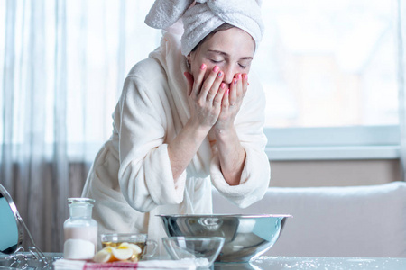 美丽的年轻女人，头上有一条毛巾，早上用水洗脸。 卫生及护理家居皮肤的概念