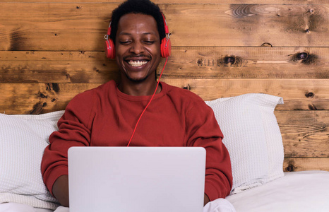 年轻的美国黑人男子使用他的笔记本电脑带耳机在床上。