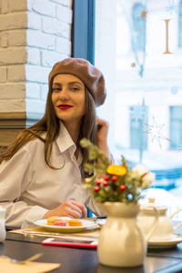 美丽的女孩坐在贝雷帽里的一张桌子旁，在一家咖啡馆里，喝着一杯茶，吃着杏仁饼干，微笑着
