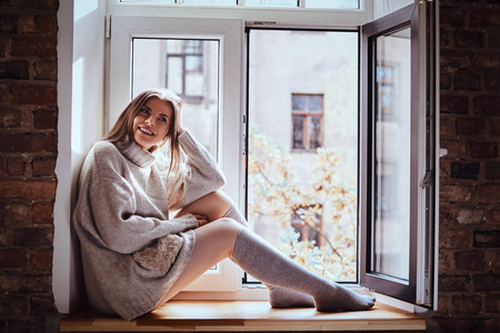 可爱的女孩穿着一件暖和的毛衣和袜子坐在窗台上, 旁边的窗户打开