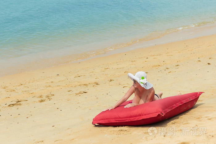 女人赤身裸体,戴着白帽子,戴着墨镜,躺在靠近水面的红色床垫上