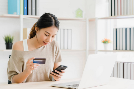 美丽的亚洲女人使用智能手机通过信用卡购买网上购物，而穿毛衣坐在家里客厅的桌子上。 生活方式女性在家的概念。