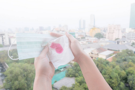 女性手或女性手拿着防护面罩和带血的餐巾纸，因为城市中的空气污染，有微粒物质或点与城市建筑背景有关