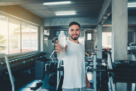 运动型男性拿着水瓶在镜头前给人拍照的照片，背景是运动器材，微笑的运动男在健身房锻炼，运动俱乐部和健康理念。