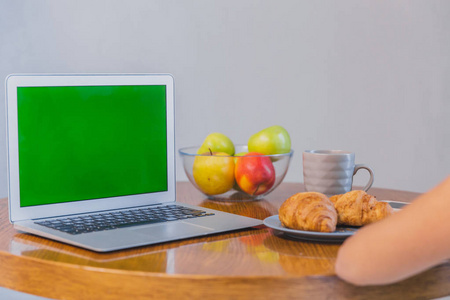 用一次性咖啡和羊角面包的女性手的裁剪快照。桌子上的笔记本电脑和配件。绿色屏幕