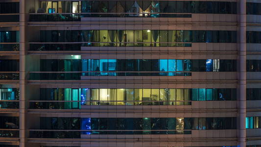 在多层现代玻璃住宅建筑中发光的窗户在夜间时间推移时亮起。 公寓里的人。 摩天大楼在晚上照明。 迪拜码头阿联酋。