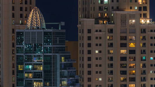 在多层现代玻璃住宅建筑中发光的窗户在夜间时间推移时亮起。 公寓里的人。 摩天大楼在晚上照明。 迪拜码头和JBR阿联酋。
