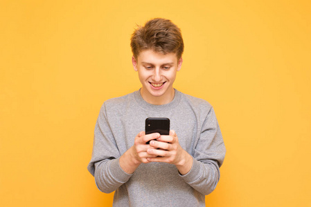 一个快乐的人的肖像，站在黄色的背景上，使用智能手机，看着屏幕。 微笑的年轻人在黄色背景下孤立的智能手机上使用互联网。