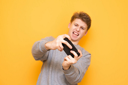 情绪紧张的家伙手里拿着手机，在黄色背景下玩游戏。 一个带着情感的年轻人正在智能手机上玩手机游戏。 手机游戏。 共用空间