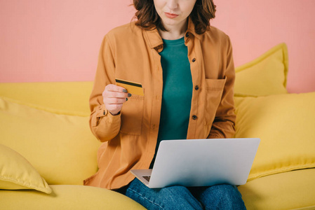 女性坐在黄色沙发上，使用笔记本电脑时持有信用卡的局部视图