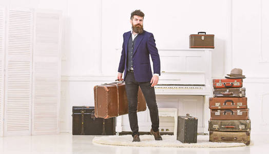 行李和度假概念。男子气概优雅, 在严格的脸上站在一堆老式手提箱, 持有手提箱。男子, 旅客与胡子和胡子的行李, 豪华白色内饰背景