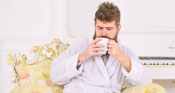 男子睡浴袍, 喝咖啡, 享受豪华酒店的香气, 在早晨, 白色背景。精英休闲理念。胡子和胡子的人享受早晨, 而坐在豪华扶手椅上