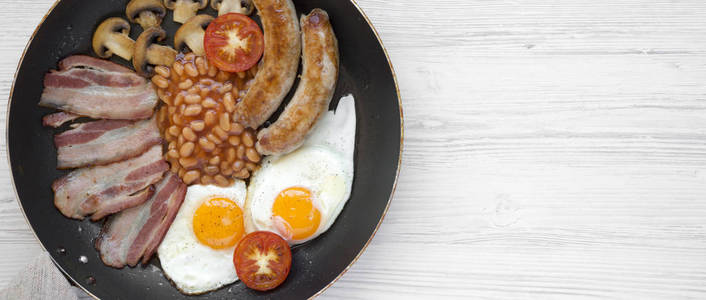 丰盛的英式早餐，在烹饪锅，香肠，煎蛋，豆子和培根，在白色的木制桌子上。 复制空间。 从上面。 平坦的。