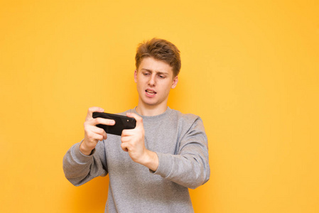 集中的家伙手里拿着智能手机，看着黄色背景上的屏幕，玩游戏。 专注的年轻人在电话上玩游戏。 移动游戏概念。