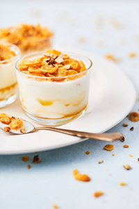 健康早餐或甜点。 美味的玉米片与酸奶杏仁和蜂蜜在一个小玻璃和一勺酸奶和玉米片在白色盘子在浅蓝色背景。 复制空间。